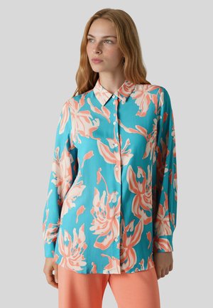 Kvinde iført en blå skjorte med store lyserøde blomsterprint og ferskenfarvede bukser, stående mod en ensfarvet grå baggrund.