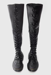 Une paire de bottes en cuir noires jusqu'aux genoux avec des semelles épaisses et des lacets sur le devant, sur un fond uni.