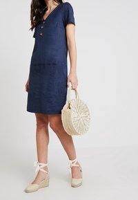 Granatowa sukienka z krótkim rękawem i dekoltem w szpic, ozdobiona trzema guzikami. W połączeniu z plecioną okrągłą torbą i beżowymi espadrylami na koturnie.