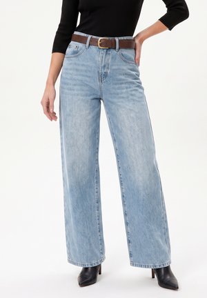 Femme portant un jean large bleu clair, des bottes à talons noires pointues, une ceinture marron et un haut à manches longues noir, debout avec une main sur la hanche.