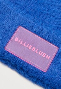 Textil azul mullido con una etiqueta rosa transparente que lleva el texto "BILLIEBLUSH" en relieve de un rosa más oscuro, cosida alrededor de los bordes.