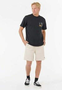 Schwarzes kurzärmliges T-Shirt mit einer kleinen gemusterten Tasche, kombiniert mit beigen Shorts. Das Modell trägt schwarze Sneaker mit weißen Schnürsenkeln und Socken.