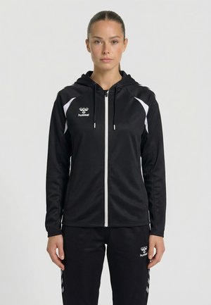 Femme debout face à l'avant portant un sweat à capuche noir zippé et un pantalon assorti avec des logos et des accents blancs Hummel sur les manches et les côtés.