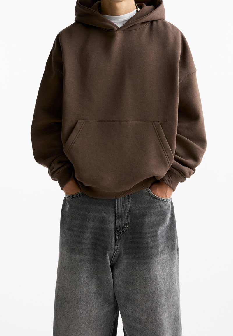 Personne portant un sweat à capuche marron avec une poche avant et un jean gris délavé et ample, les mains dans les poches.