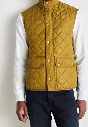 Man wearing mustard yellow quilted vest with snap buttons over white long-sleeve shirt and dark pants, standing against plain background.