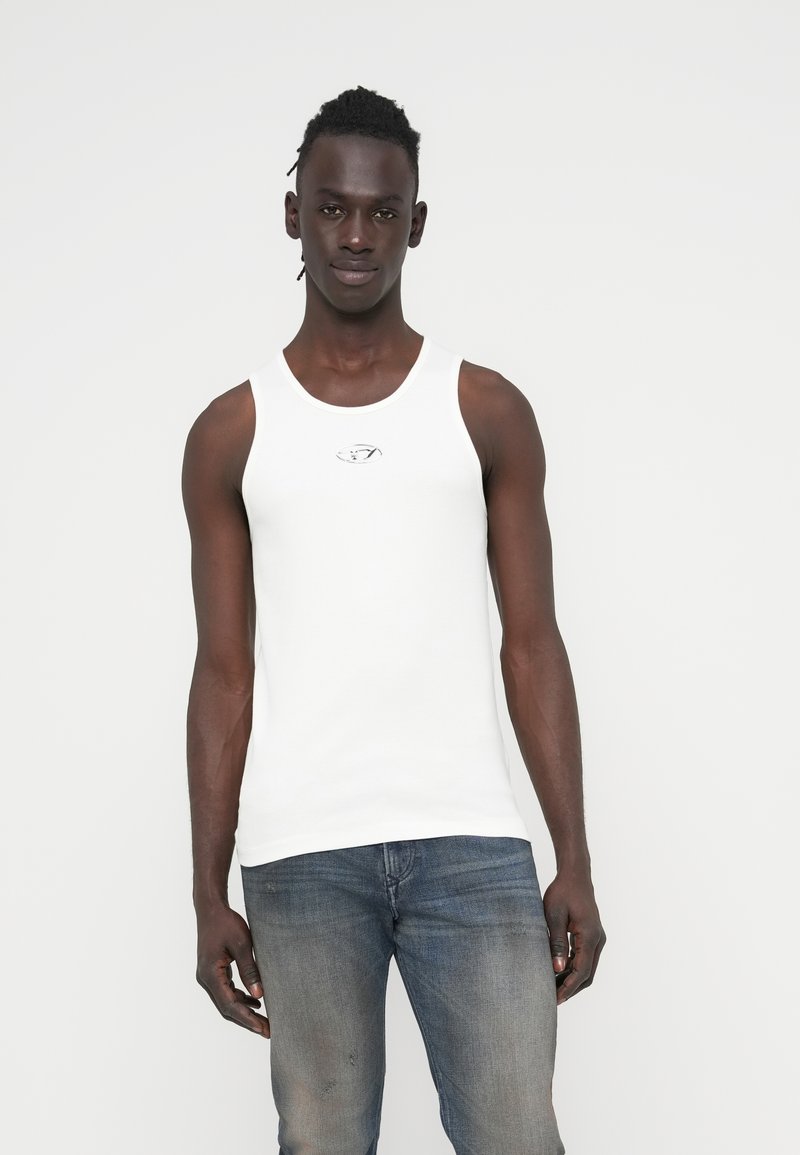 Άσπλο αμάνικο τοπ με στρογγυλή λαιμόκοψη και ασημένιο λογότυπο στο στήθος, σε συνδυασμό με σκούρο μπλε τζιν παντελόνι.