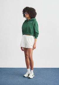 Hoodie vert court, short de survêtement blanc et baskets blanches avec des accents verts. Cheveux épais et bouclés, et lunettes de soleil rondes violettes complètent le look.