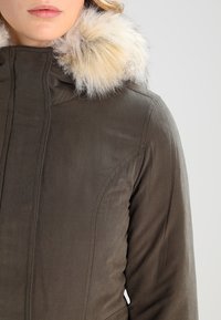 Kurtka w kolorze oliwkowej zieleni z zamkiem błyskawicznym z przodu i futrzanym kołnierzem z imitacji futra. Charakteryzuje się przeszyciami oraz gładką fakturą.