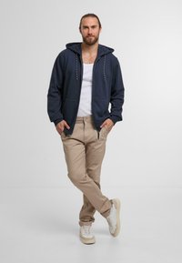 Navy zip-up hoodie with drawstrings, light tank top, beige pants, and white sneakers with beige accents, standing against a neutral background.