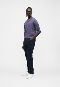 Maglione viola in cotone con scollo a V e piccolo logo giallo, abbinato a pantaloni blu navy e sneakers bianche. Design semplice, vestibilità comoda.