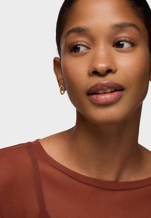 Giovane donna con pelle liscia che indossa piccoli orecchini a cerchio in oro e un top color ruggine, che guarda leggermente a destra su uno sfondo bianco.