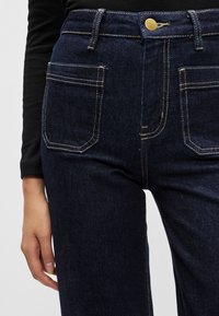 Magas derekú sötét farmernadrág kontrasztos sárga öltésekkel, két elülső zsebbel és egy arany gombbal a derékrészen.