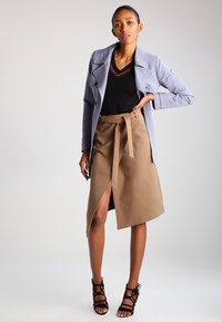 Manteau bleu clair à double boutonnage porté sur un haut noir, associé à une jupe portefeuille marron avec une fente latérale et une ceinture nouée. Sandales à talons noires.