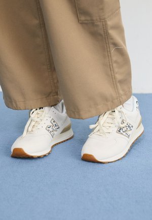 Baskets blanches avec des accents imprimé léopard portées avec un pantalon beige large sur un sol recouvert d'un tapis bleu clair.