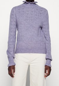 Ljuslila stickad tröja med hög rynkad halsringning, med intrikata spetsmönster på ärmarna och kroppen. Texturerad yta.