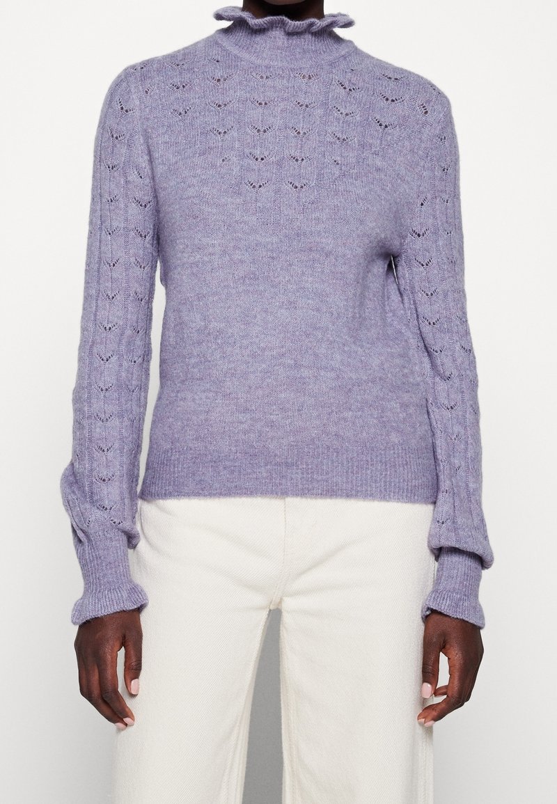 Ljuslila stickad tröja med hög rynkad halsringning, med intrikata spetsmönster på ärmarna och kroppen. Texturerad yta.