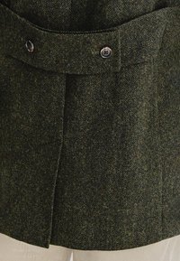 Zielona wełniana kurtka w wzór jodełki z paskiem, z podwójnymi guzikami i usztywnianym kołnierzem, o teksturowanym wykończeniu.