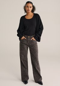 Cardigan nero con vestibilità morbida, indossato sopra una maglia nera. Paio di jeans marroni con stampa leopardata, vita alta e tasche frontali. Scarpe nere.