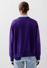 Paarse sweatshirt met een relaxed fit en geribbelde manchetten, gedragen over een lichtblauw overhemd met kraag. Zichtbare denim patch op de broek.