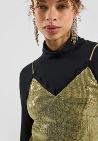 Frau mit lockigem Haar, die lange, hängende Kristallohrringe, einen schwarzen Rollkragenpullover und ein goldenes Camisole mit Pailletten und Spitzenbesatz trägt.