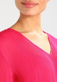 Fuchsiový top s vertikálními sklady, výstřihem do V, hladkou strukturou a přiléhavým střihem, který zdůrazňuje detaily na ramenou a výstřihu.