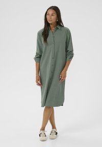 Robe boutonnée en tissu vert doux, mi-longue, avec des manches retroussables, présentant une coupe droite et une texture subtile. À associer avec des baskets.