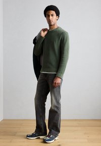 Zeleni rebrasti pulover, sivi jeans in večbarvni športni copati. Model drži jakno čez eno ramo. Gladko leseno tla.