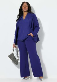 Traje a medida púrpura con una parte superior holgada y pantalones de pierna ancha. Tacones plateados y un bolso con patrón completan el conjunto. Tejido suave y ligero.