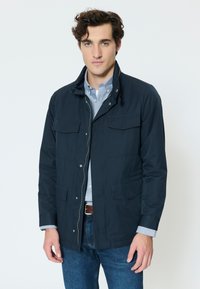 Young man with curly hair wearing a navy jacket over a light blue button-up shirt and blue jeans, standing against a plain white background.