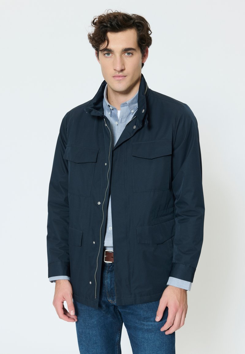 Young man with curly hair wearing a navy jacket over a light blue button-up shirt and blue jeans, standing against a plain white background.