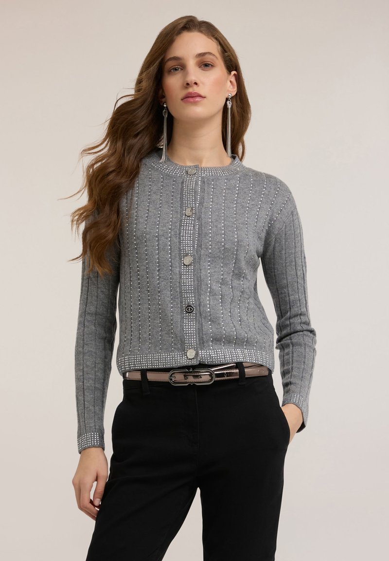Szary prążkowany kardigan z okrągłym dekoltem, ozdobiony guzikami i srebrnymi detalami na dole, noszony na czarnych spodniach.