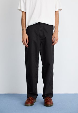 Black cotton trousers feature a straight-leg design with a button closure and side pockets, paired with brown leather shoes.