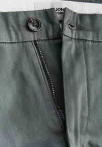 Gros plan sur la fermeture éclair d'un pantalon gris partiellement ouverte avec un bouton noir fixé et une boutonnière visible sur la ceinture.