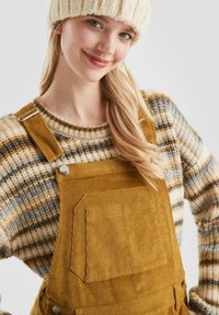 Mosterdkleurige corduroy tuinbroek met een voorzak, gedragen over een meerkleurige, gestreepte gebreide trui. Model heeft lang haar en een crèmekleurige beanie.