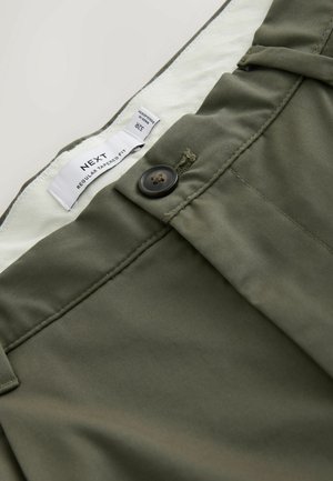 Olivgrüne, schmal zulaufende Hose mit schwarzem Knopf und sichtbarem weißem Innenschild mit der Aufschrift „NEXT Regular Tapered Fit 32R.“