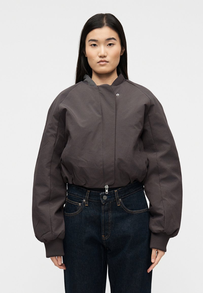 Γυναίκα που φοράει ένα σκούρο γκρι φουσκωτό bomber μπουφάν με φερμουάρ και κουμπί, σε συνδυασμό με ψηλόμεσα σκούρα τζιν, στέκεται ίσια.