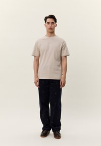 Fiatal férfi áll egyszínű háttér előtt, bézs rövid ujjú inget, sötét mintás nadrágot és barna cipőt visel, előre néz.