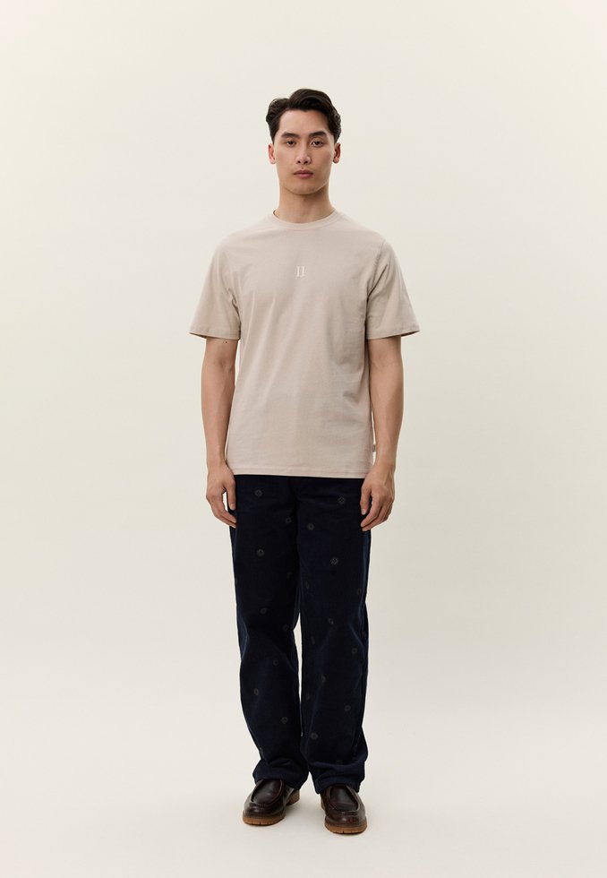 Fiatal férfi áll egyszínű háttér előtt, bézs rövid ujjú inget, sötét mintás nadrágot és barna cipőt visel, előre néz.