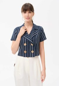 Granatowa krótka podwójnie zapięta kurtka w białe pionowe paski, z krótkimi rękawami i czterema drewnianymi guzikami z przodu, w połączeniu z jasnymi spodniami.