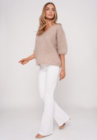 Beiger, kuscheliger V-Ausschnitt-Pullover mit Puffärmeln, kombiniert mit weißen Schlagjeans und nudefarbenen Sandalen, vor einem schlichten weißen Hintergrund.