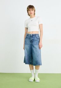 Hvit avkortet t-skjorte med svart logo, blå denim culotte shorts med en falmet design, kombinert med neon grønne sandaler og hvite sokker.
