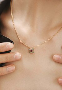 Collar de cadena de oro con un colgante cuadrado multifacético que presenta acentos en rosa y azul, colocado sobre un tono de piel neutro.