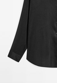 Camicia nera a maniche lunghe con una texture morbida, orlo arrotondato e chiusura con bottoni. I polsini presentano una finitura a contrasto.