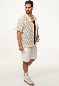 Beige offen gestricktes kurzärmliges Hemd, schwarzes Top, weiße Shorts und cremefarbene Sneakers mit braunen Akzenten. Dazu gehören grün- und gelb gestreifte Socken.