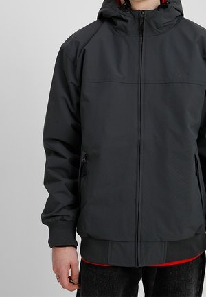 Person trägt eine schwarze Kapuzenjacke mit Reißverschluss, seitlichen Taschen und gerippten Bündchen, teilweise über einem roten Shirt und dunklen Hosen.