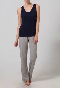 Mulher vestindo uma blusa azul-marinho sem mangas e calças soltas cinza-claro, de pé descalça contra um fundo liso claro.