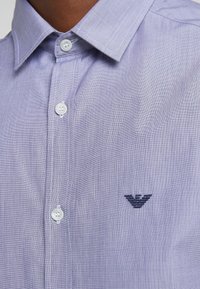 Chemise unie mauve clair à col structuré, avec des boutons blancs et un logo brodé bleu marine. Le tissu a une finition légèrement texturée.
