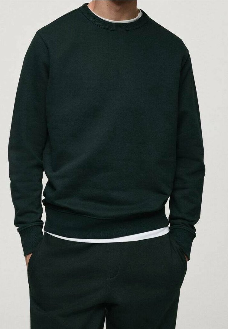 Ciemnozielony sweter z okrągłym dekoltem, z ściągaczami przy mankietach i dolnym obszyciu. Miękka tkanina, luźny krój. Nałożony na biały shirt w okolicy dekoltu.