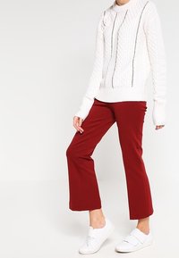 Weißer Pullover mit Zopfstrickmuster und rundem Halsausschnitt, kombiniert mit burgunderfarbenen, verkürzten Hosen und weißen Slip-On-Sneakers.