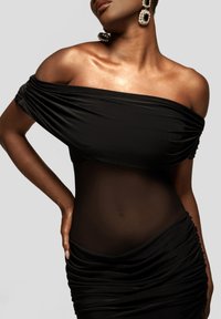 Musta olkapäät paljastava mekko, jossa on rypytettyjä yksityiskohtia, läpinäkyvä vyötäröosa ja sileä pinta, korostettuna suurilla, pyöreillä korvakoruilla.